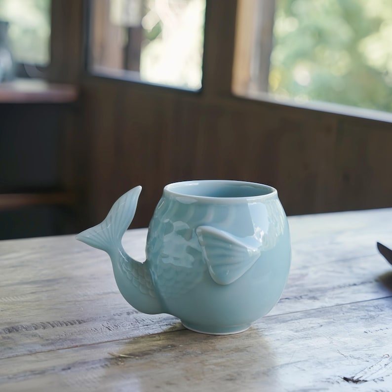 May include: A light blue ceramic mug shaped like a whale, with a tail handle and a fin on the side. The mug has a textured surface and sits on a wooden table. The mug is designed for beverages.