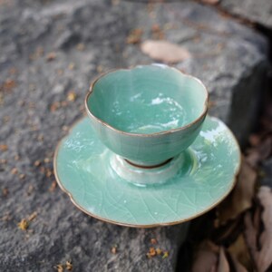 May include: A light mint green ceramic tea cup and matching saucer set featuring a delicate scalloped edge design and a visible crackle glaze finish. The cup has a rounded bowl shape and sits centered on the circular saucer.