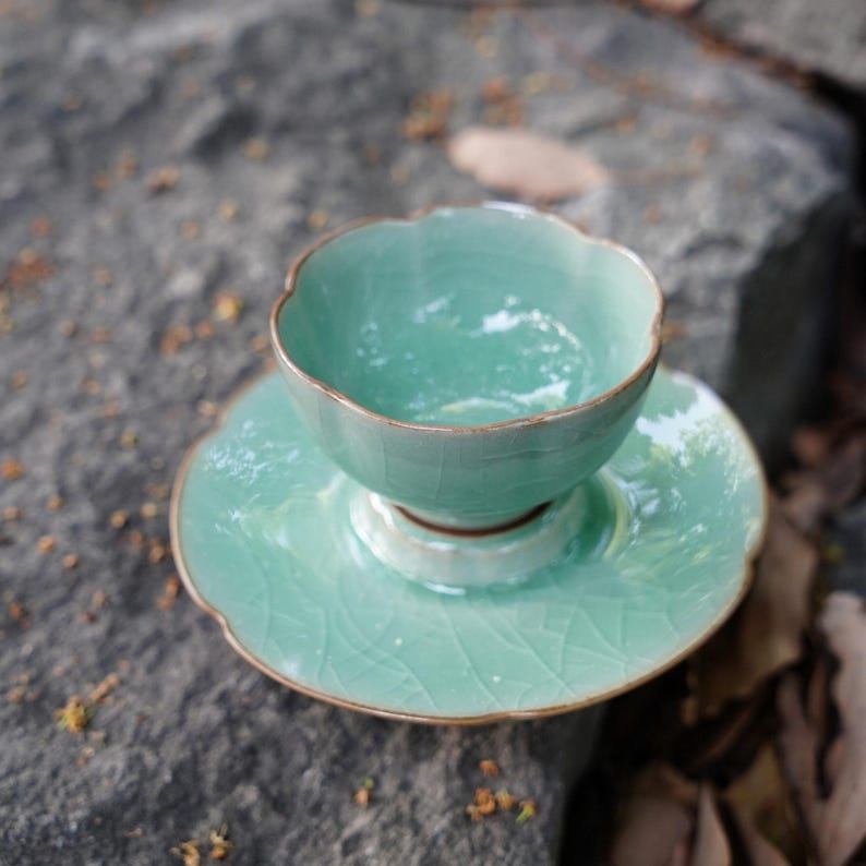 May include: A light mint green ceramic tea cup and matching saucer set featuring a delicate scalloped edge design and a visible crackle glaze finish. The cup has a rounded bowl shape and sits centered on the circular saucer.