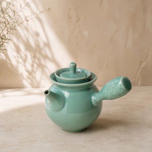 Może przedstawiać: Turkusowy ceramiczny imbryk z okrągłym korpusem, małym dziobkiem i bocznym uchwytem. Pokrywka spoczywa na imbryku, a uchwyt ma dekoracyjny kwiatowy wzór. Imbryk jest w jasnym odcieniu turkusu i ma błyszczące wykończenie.