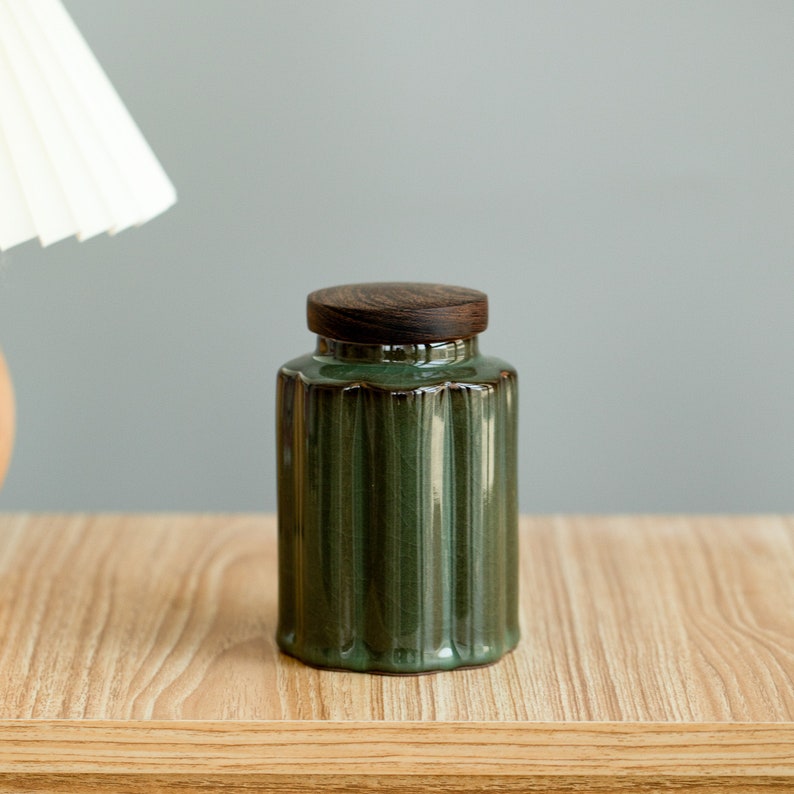 May include: A green ceramic jar with a ribbed design and a dark wooden lid. The jar is cylindrical and sits on a wooden surface. The lid is a dark brown color. The jar is approximately 5 inches tall.
