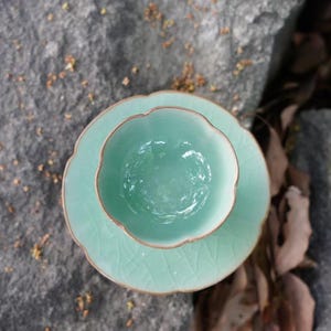 May include: A light mint green ceramic tea cup and matching saucer set. The cup features a scalloped rim and a smooth interior, while the saucer displays a delicate crackle glaze pattern across its surface, creating a cohesive look.