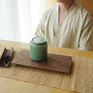 May include: A light green ceramic tea caddy with a floral design and a matching lid sits on a carved wooden tray. A wooden scoop with tea leaves is to the left. The scene is set on a table with a neutral-toned runner.