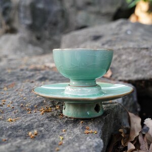 May include: A light green ceramic tea cup with a crackle glaze finish sits atop a matching saucer and a pedestal base. The cup features a scalloped rim, while the base has small cutouts, creating a cohesive set with a glossy texture.