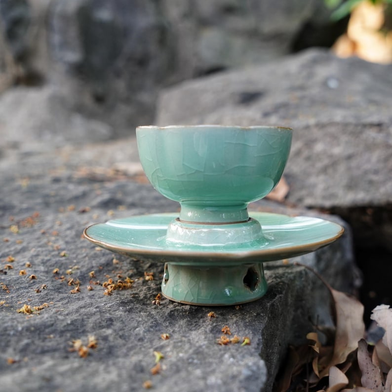 May include: A light green ceramic tea cup with a crackle glaze finish sits atop a matching saucer and a pedestal base. The cup features a scalloped rim, while the base has small cutouts, creating a cohesive set with a glossy texture.