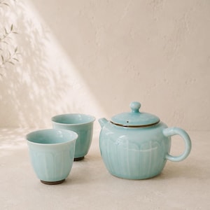 May include: A light blue ceramic teapot with a matching lid and two small matching cups. The teapot and cups feature a vertical ribbed texture and a smooth, glossy finish. The teapot has a curved handle and a small spout, all in a consistent pale blue.