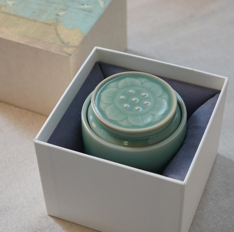 May include: A light teal ceramic jar with a floral design on the lid, nestled inside a white box with a blue-gray lining. The lid features a raised flower pattern with small circular indentations. The box is partially open, revealing the jar.