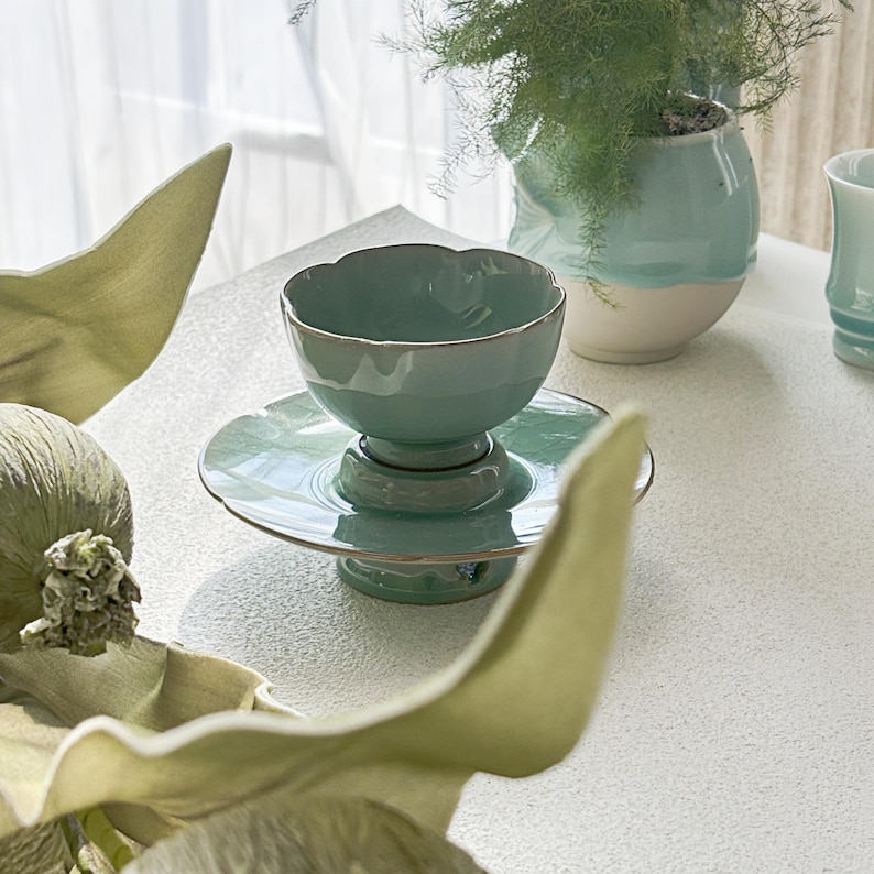 May include: A light green ceramic bowl with a scalloped rim sits atop a matching pedestal saucer. The bowl and saucer feature a glossy finish with a subtle metallic-toned edge. The set is positioned on a textured white surface.