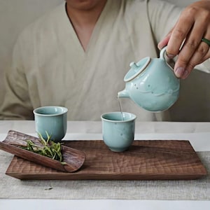 May include: A tea set with a light blue ceramic teapot pouring into a matching cup. Two cups and a wooden tray with tea leaves are also present. The tray is made of dark wood.