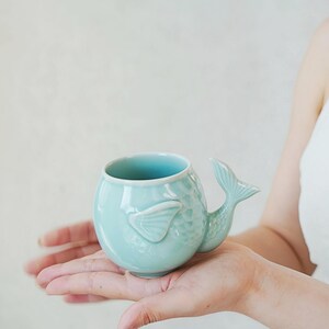 May include: A light blue ceramic mug shaped like a fish, with a tail handle and fin details. The mug has a rounded body and a smooth, glossy finish. The mug is held in a person's hands.