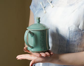 May include: A teal ceramic teacup with a lid and a handle. The teacup has a decorative design on the side.