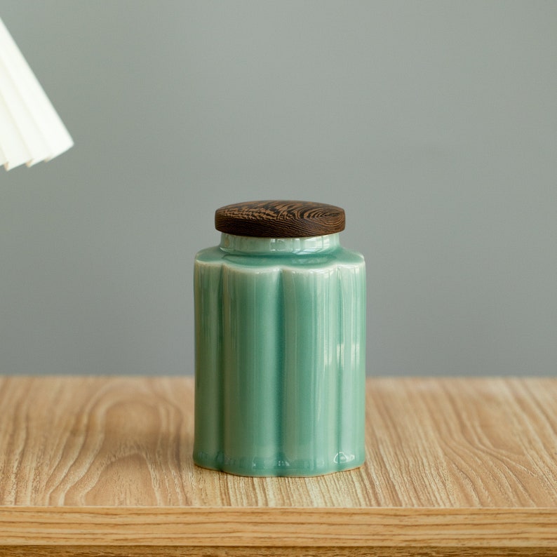 May include: A teal ceramic jar with a wooden lid. The jar has a ribbed design and a scalloped base. The lid is a dark brown color with a wood grain pattern. The jar is sitting on a light wood surface.