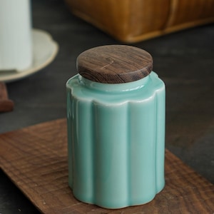 May include: A small, turquoise ceramic jar with a scalloped rim and a wooden lid. The jar has vertical ribbed detailing and sits on a wooden tray. The lid is a dark brown color with a wood grain pattern. The jar is a decorative item.