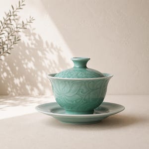May include: A light green ceramic gaiwan tea set consisting of a lidded bowl and a matching saucer. The exterior of the bowl and the lid feature a raised, embossed floral pattern with a glossy finish, showcasing a consistent celadon-toned glaze.