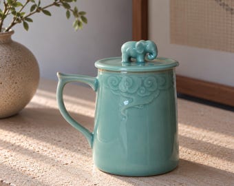 Könnte beinhalten: Ein hellblauer Keramikbecher mit passendem Deckel, der eine kleine Elefantenfigur als Griff trägt. Der Becher hat einen Henkel und ein Reliefmuster aus stilisierten Wolken im oberen Bereich, überzogen mit einer glatten, glänzenden Glasur.