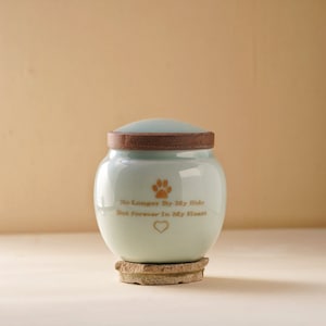 May include: A light blue ceramic pet urn with a wooden lid. The urn has a gold paw print and the text "No Longer By My Side But Forever In My Heart" with a gold heart.
