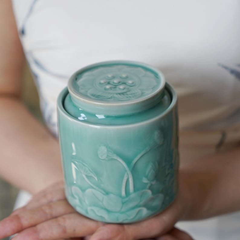 May include: A teal ceramic jar with a lid, featuring embossed lotus flower designs. The jar is cylindrical with a slightly rounded lid. The lotus flower design is repeated on the lid. The jar is held in a person's hands.