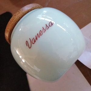 May include: A light blue ceramic jar with a wooden lid. The jar has the name "Vanessa" written in red cursive on the front.