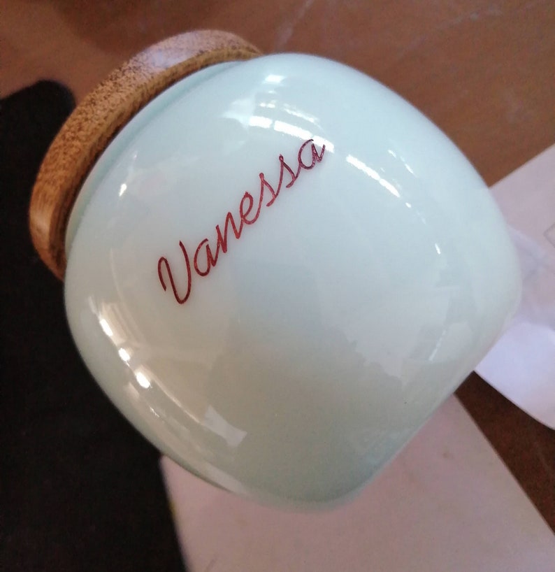 May include: A light blue ceramic jar with a wooden lid. The jar has the name "Vanessa" written in red cursive on the front.