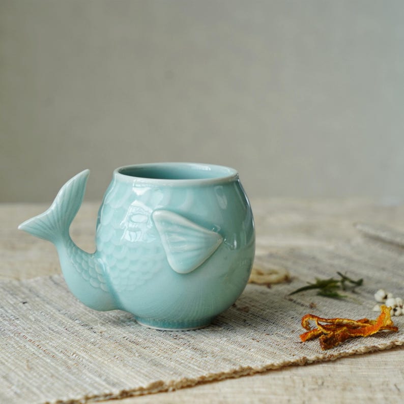 May include: A light blue ceramic mug shaped like a fish, featuring a tail and fin details. The mug has a textured body and a smooth, glossy finish. The mug is sitting on a neutral-colored surface.