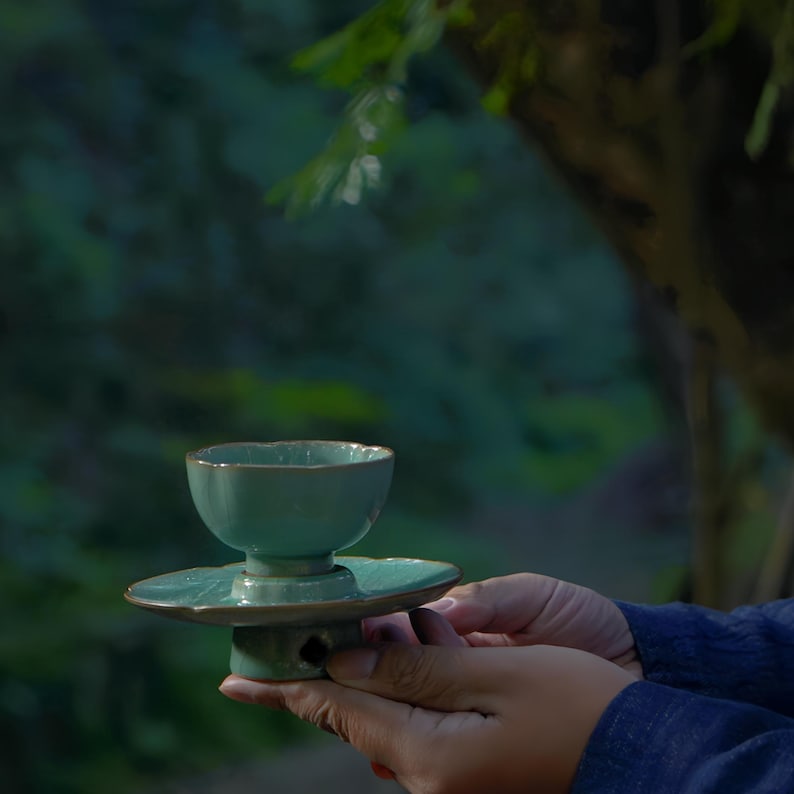 May include: A light green ceramic tea cup with a matching saucer, featuring a scalloped rim design and a pedestal base. The glaze has a subtle crackle texture, and the cup sits securely on the circular saucer, which rests on a small base.