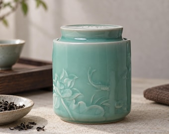 May include: A cylindrical celadon green ceramic tea canister featuring a relief carving of a lotus flower and a swimming fish on its exterior. The container includes a matching fitted lid with a smooth, glossy finish and subtle crackle glaze.