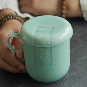 May include: A light teal ceramic mug with a matching lid. The mug has a curved handle and a rounded body. The lid and the mug have a rectangular design with a wave pattern. The mug is held by a person.