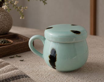 May include: A light blue ceramic mug with a matching lid, featuring a rounded body and a single handle. The surface is decorated with dark brown, irregular, organic-shaped spots and streaks distributed across the lid and the mug body.