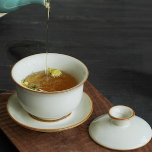 May include: A white ceramic tea cup and saucer set with a lid, featuring a brown trim. The cup is filled with tea and flower buds, with tea being poured from a teal teapot. The set is on a wooden surface.