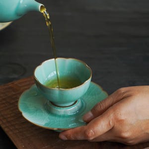May include: A light turquoise ceramic tea cup and matching saucer with a delicate crackle glaze finish. The cup features a scalloped rim and is being filled with golden tea poured from a matching teapot, showcasing a refined aesthetic.