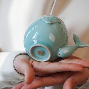 May include: A light blue ceramic whale-shaped tea cup. The cup features a detailed tail, a spout, and a shell-like design on top. White spots accent the whale's body. The cup is held in a person's hands.
