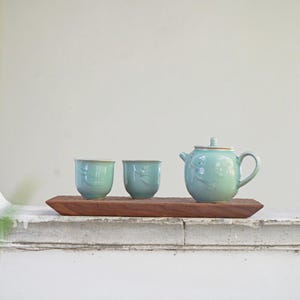 May include: A light blue ceramic tea set with two cups and a teapot, presented on a rectangular wooden tray. The teapot and cups feature a floral design. The tea set is placed on a white surface.