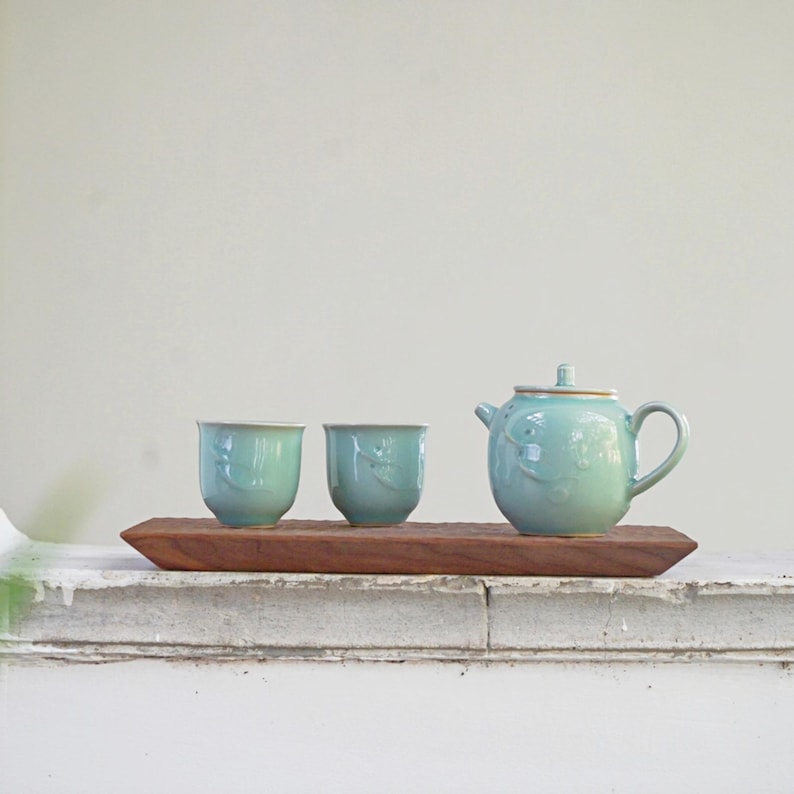 May include: A light blue ceramic tea set with two cups and a teapot, presented on a rectangular wooden tray. The teapot and cups feature a floral design. The tea set is placed on a white surface.