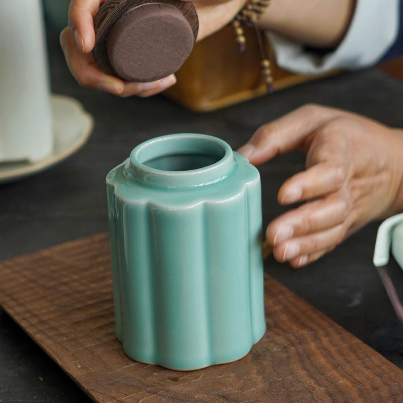 May include: A light teal ceramic jar with a fluted design and a wooden lid. The jar is cylindrical with a scalloped top and sits on a wooden surface. The lid is being held above the jar.