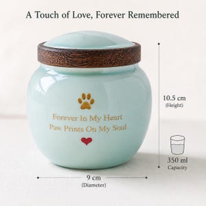 Peut inclure: Une urne en c&eacute;ramique bleu clair avec un couvercle en bois fonc&eacute;. Le texte indique A Touch of Love, Forever Remembered et Forever In My Heart Paw Prints On My Soul, avec une patte dor&eacute;e et un c&oelig;ur rouge. Hauteur 10,5 cm, diam&egrave;tre 9 cm, capacit&eacute; 350 ml.