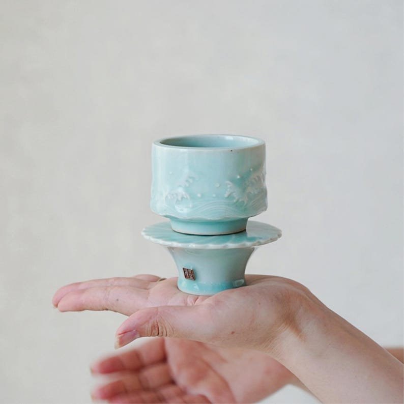 May include: A light blue ceramic teacup and saucer set. The cup features a wave design around the rim. The set is held in a hand, showcasing the delicate craftsmanship and the soft, pastel color of the ceramic.