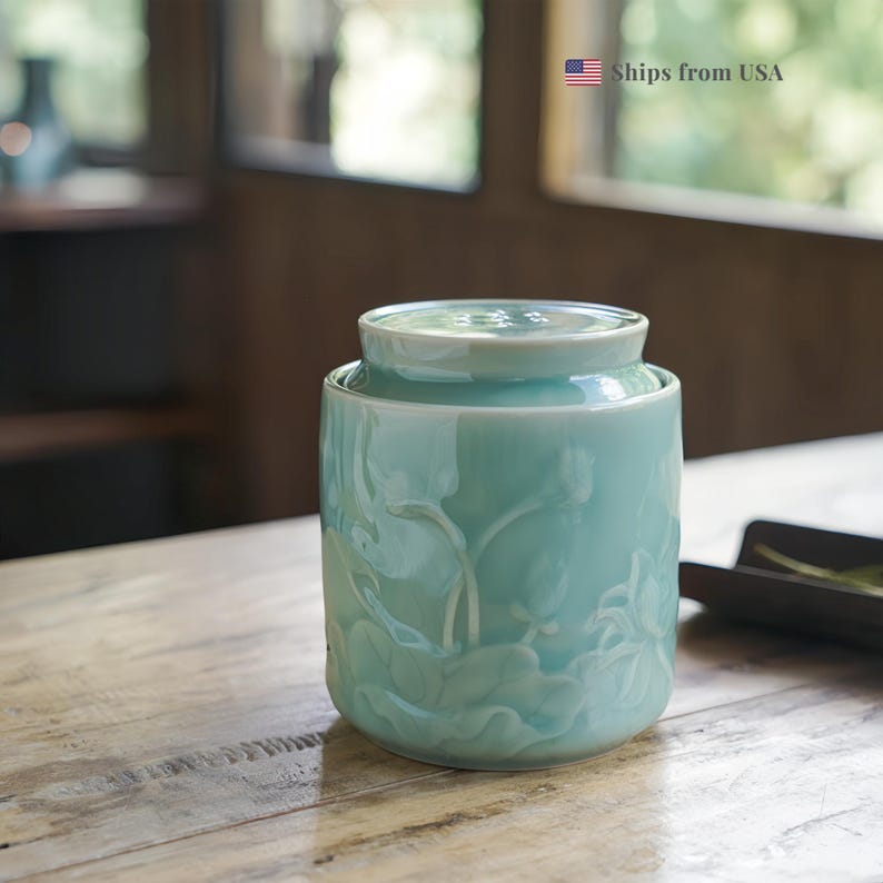 May include: A light teal ceramic jar with a lid, featuring embossed lotus flower and leaf designs. The jar is cylindrical with a slightly rounded top and a matching lid. The background is blurred, suggesting a natural setting.