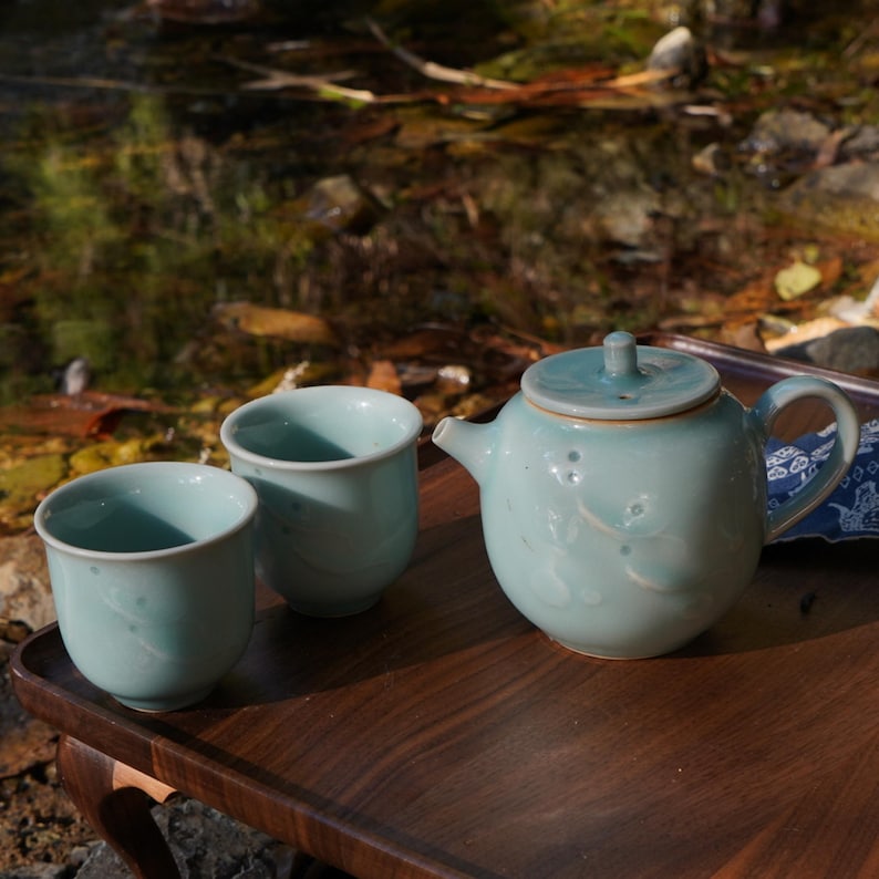 May include: A pale blue ceramic tea set comprising two cups and a teapot with a lid. The teapot and cups feature a subtle floral pattern. The set is presented on a dark wooden tray.