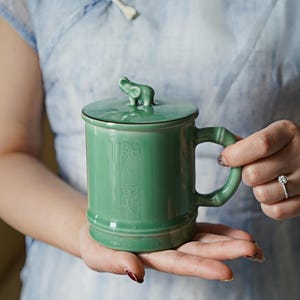 May include: A green ceramic mug with a lid featuring a small elephant figurine. The mug has a bamboo-style handle and a design of Chinese characters on the side.