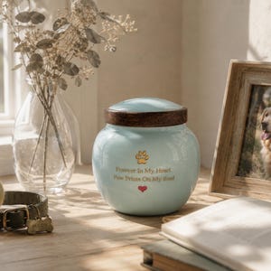 Peut inclure: Une urne pour animal de compagnie en c&eacute;ramique bleu clair avec un couvercle en bois brun fonc&eacute;. L'avant pr&eacute;sente une empreinte de patte dor&eacute;e, un petit c&oelig;ur rouge et le texte dor&eacute; : Forever In My Heart, Paw Prints On My Soul. Forme arrondie.