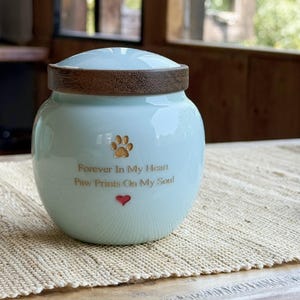Peut inclure: Urne funéraire pour animaux de compagnie en céramique bleu clair, avec un couvercle en bois. Elle présente une empreinte de patte et un cœur, avec le texte "Forever In My Heart Paw Prints On My Soul".