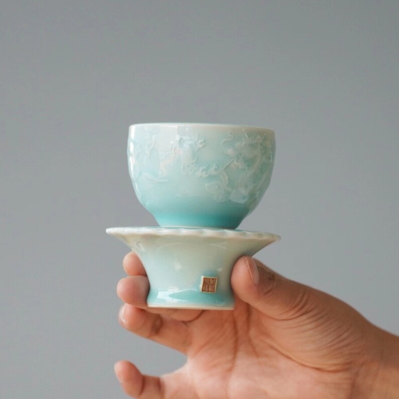 May include: A light blue ceramic teacup and saucer set. The teacup features a floral design and sits atop a matching saucer. The set is held in a hand against a gray background. The teacup and saucer are made of ceramic.