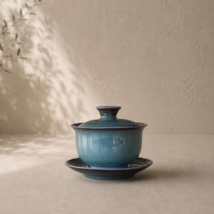 May include: A teal ceramic gaiwan tea set with a matching lid and saucer. The gaiwan has a textured surface and a dark purple rim. The set is designed for traditional tea ceremonies.