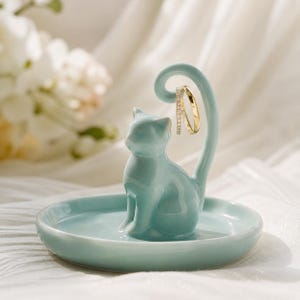 May include: A light blue ceramic cat-shaped ring holder. The cat sits on a round dish with its tail curved upwards, holding two rings. The rings are gold and silver. The holder is on a white surface.