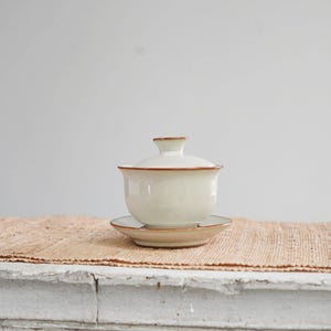 Op de afbeelding: Een witte keramische gaiwan theeset met een bijpassende deksel en schotel. De randen van de gaiwan en het deksel zijn afgewerkt met een bruin accent. De set wordt gepresenteerd op een beige geweven mat.