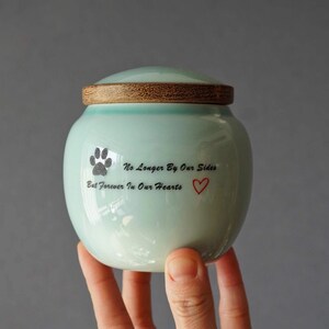 May include: A light blue ceramic pet urn with a wooden lid. The urn has a paw print and the text "No Longer By Our Sides But Forever In Our Hearts" with a red heart.