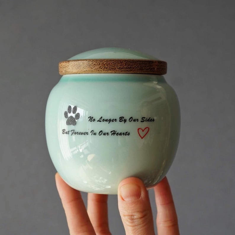 May include: A light blue ceramic pet urn with a wooden lid. The urn has a paw print and the text "No Longer By Our Sides But Forever In Our Hearts" with a red heart.