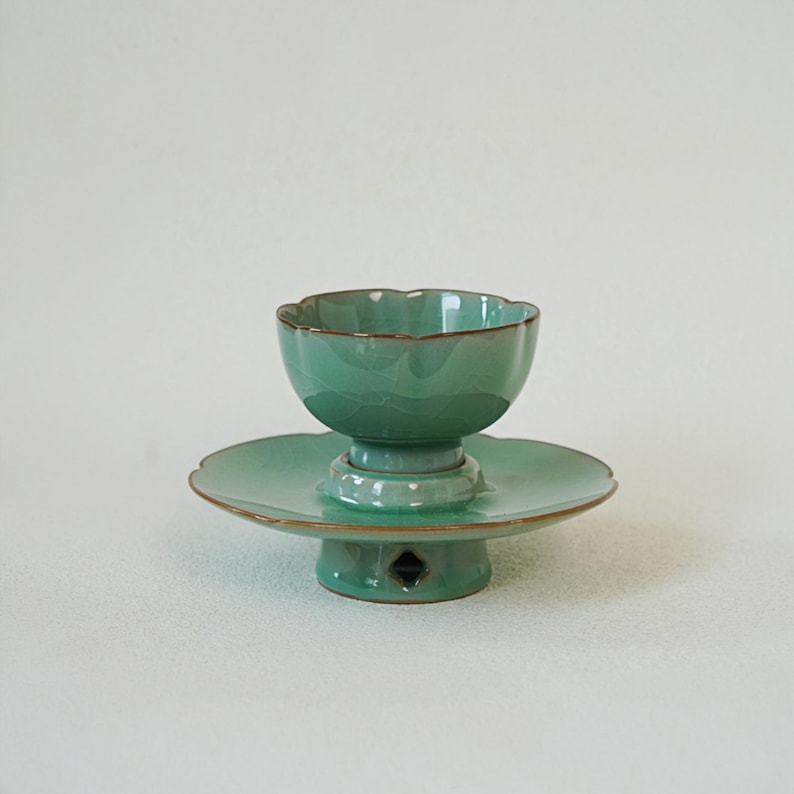 May include: A celadon green ceramic tea bowl with a scalloped rim, resting on a matching pedestal saucer. The saucer features a wide, flat base with a small diamond-shaped cutout. The glaze has a subtle crackle finish and brown edges.