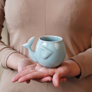 May include: A light blue ceramic mug shaped like a whale, featuring a tail handle and fin details. The mug is held in open hands, showcasing its unique design. The mug is a light blue color.