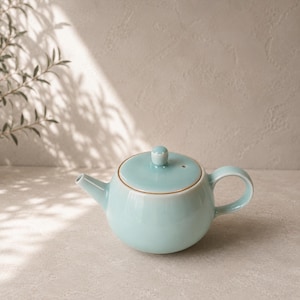 Peut inclure: Une théière en céramique bleu clair avec un corps arrondi, une anse incurvée et un bec verseur. Le couvercle a un petit bouton. La théière est posée sur une surface claire, avec des ombres d'une plante en arrière-plan.